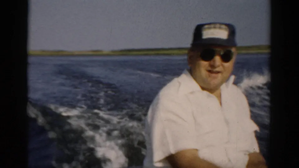 Continued shot of man in white shirt and sunglasses at stern of moving speedboat, shoreline with trees, 1957 home movie, Catskill area.