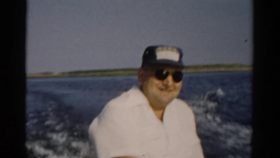 Final frame of man in white shirt and sunglasses smiling at camera from speedboat stern, 1957 home movie, possibly Catskill area, New York.