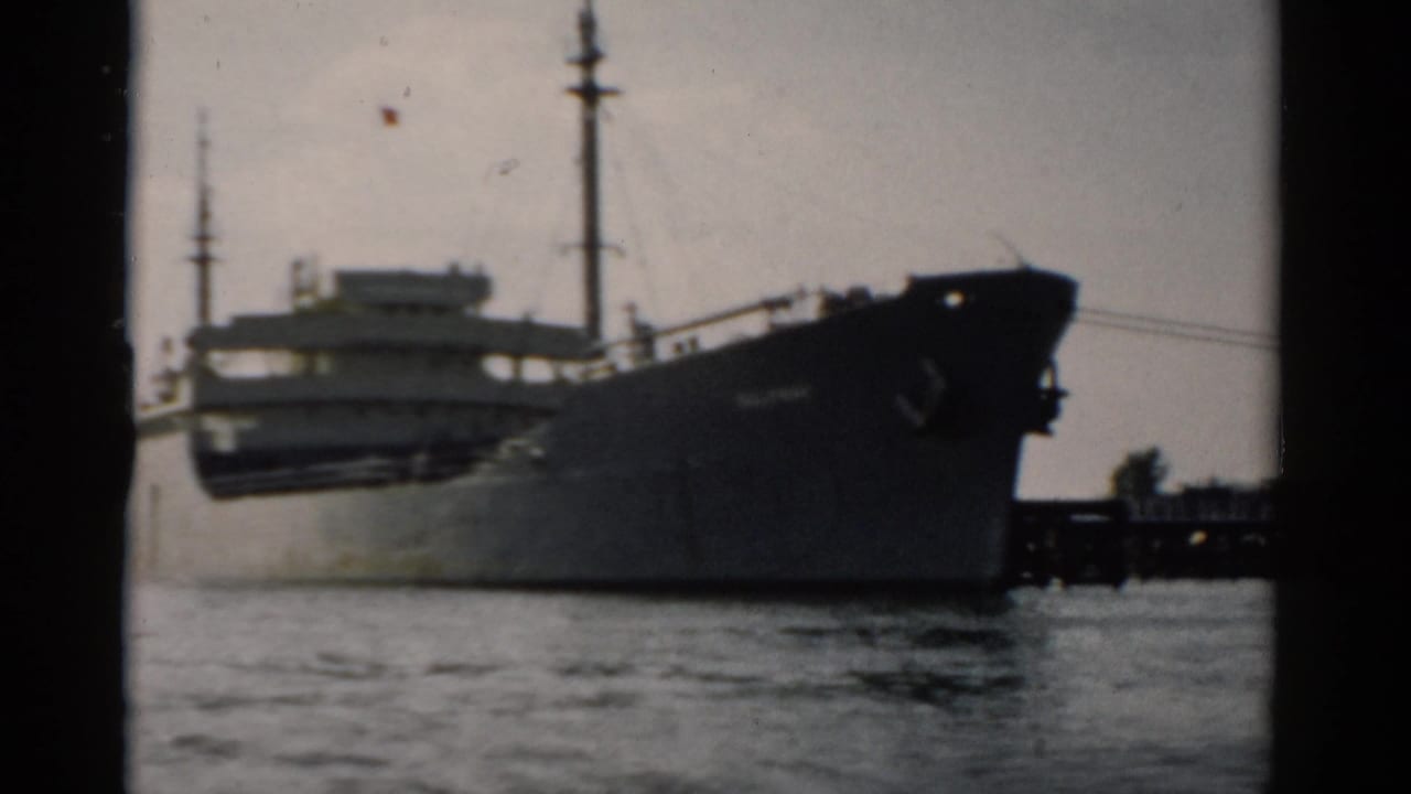 Large dark-hulled cargo freighter passing on calm water, shot on 8mm film circa 1957, possibly the Hudson River near Catskill, New York.