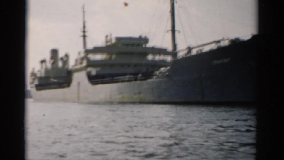 Large dark-hulled cargo vessel moored or passing alongside a concrete quay wall, 8mm home-movie footage circa 1957, possibly the Hudson River near Catskill, New York.