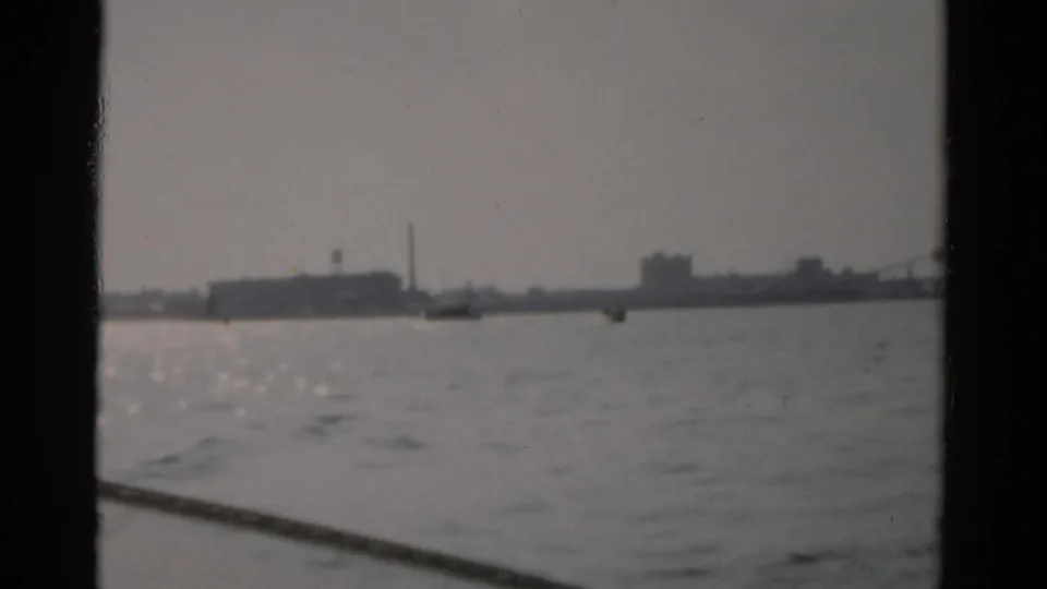 Wide 8mm shot across calm water toward industrial waterfront, circa 1957, possibly near Catskill, NY; hazy flat light, buildings on horizon.