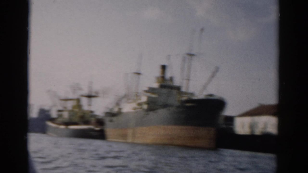 A large mid-century cargo ship with derrick cranes at a harbor dock, filmed from a moving boat on what metadata identifies as a New York waterway, circa 1957.