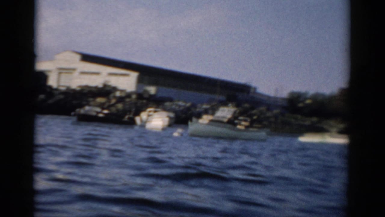 An industrial harbor shed with moored small vessels on a northeastern U.S. waterfront, filmed from a moving boat in 8mm color, circa 1957.