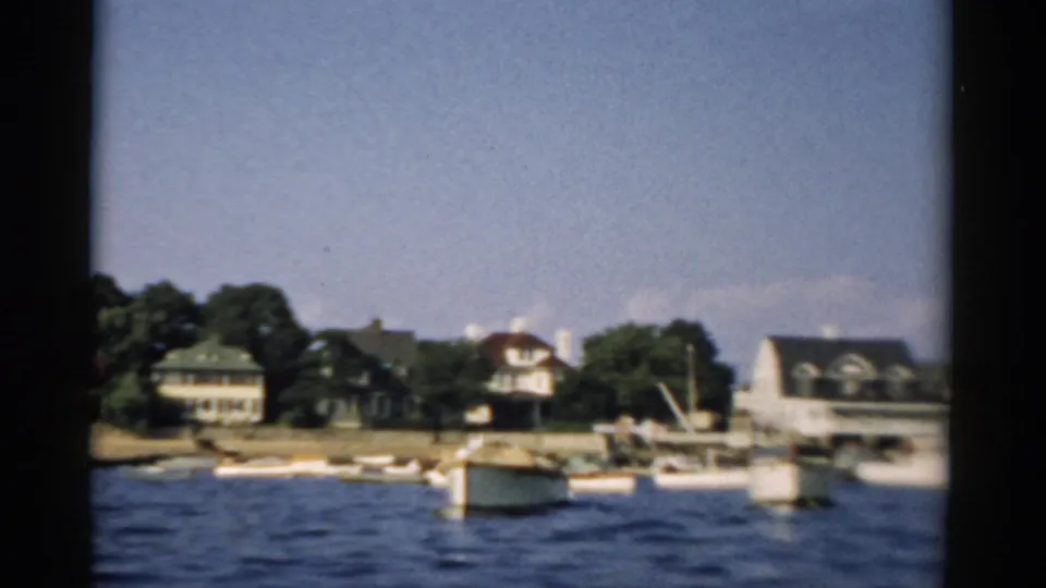 A small-town northeastern U.S. waterfront showing residential buildings, deciduous trees, and moored small craft, filmed from a moving boat circa 1957.