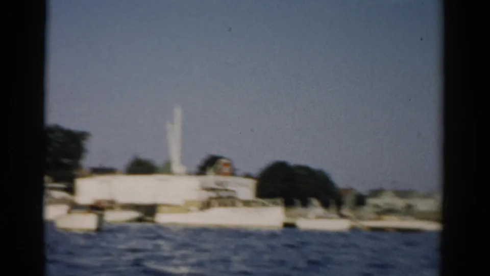 A large white building — possibly industrial or institutional — with a tall structure (smokestack or antenna) on a northeastern U.S. shoreline, filmed in 8mm color circa 1957.
