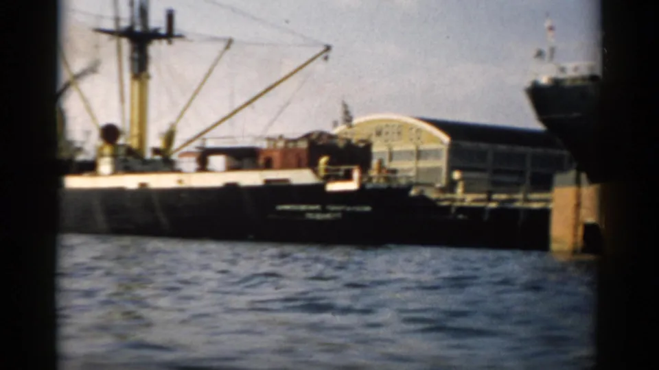 Cargo vessel or barge with mast rigging alongside dock, Catskill NY, c. 1957 — curved-roof waterfront building visible behind hull, 8mm color.