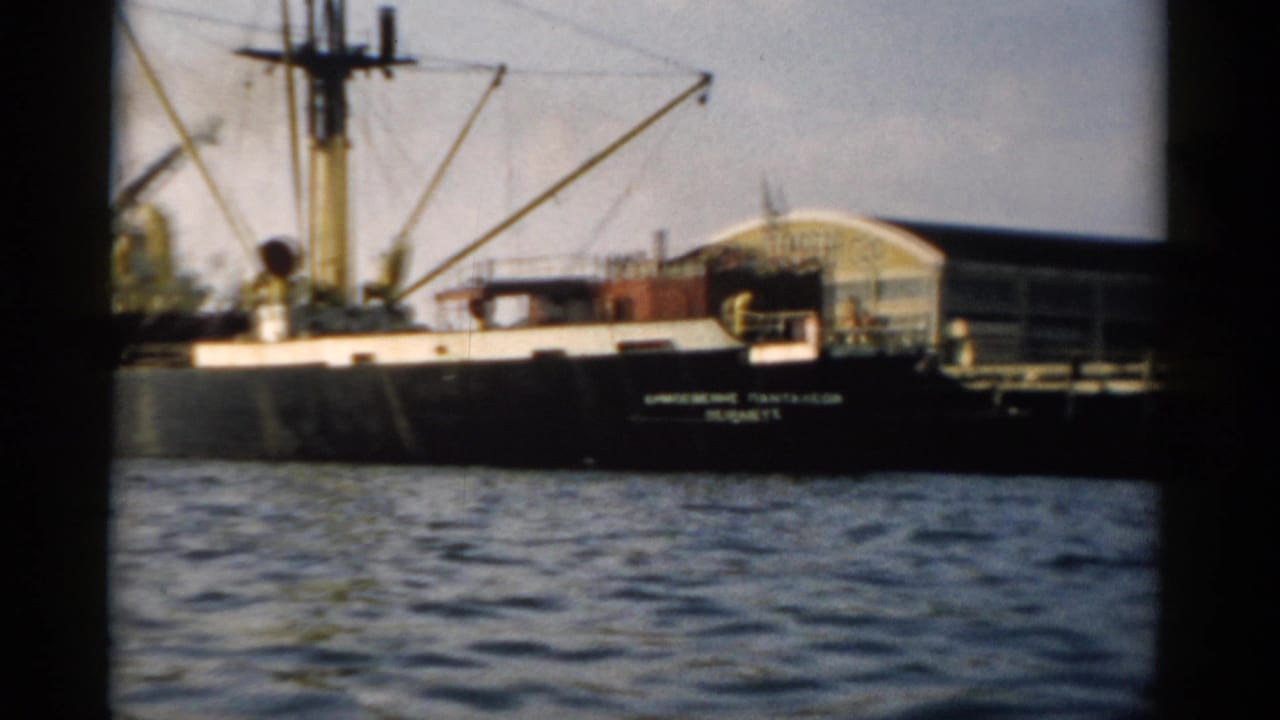 Black-hulled cargo ship with derrick cranes moored at what appears to be a Hudson River port, circa 1957, captured on 8mm film.