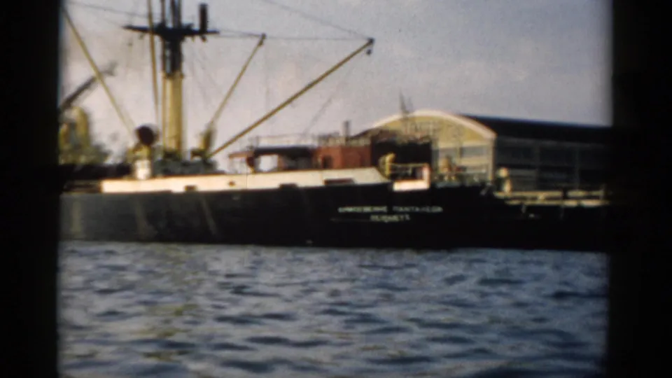Black-hulled cargo ship with derrick cranes moored at what appears to be a Hudson River port, circa 1957, captured on 8mm film.
