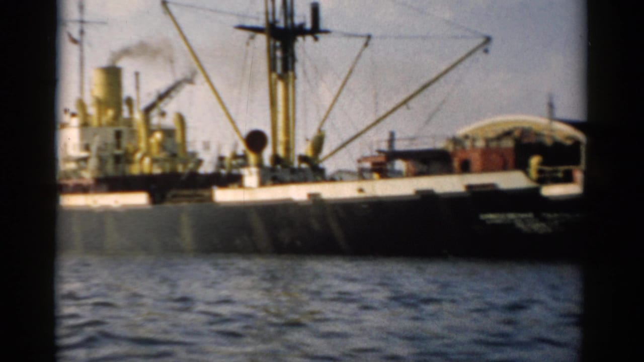 Cargo ship with yellow derrick masts and active smokestack photographed from a passing pleasure boat, appearing to show a Hudson River port scene circa 1957.