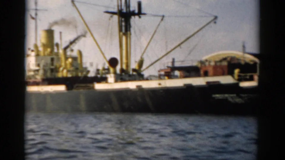 Cargo ship with yellow derrick masts and active smokestack photographed from a passing pleasure boat, appearing to show a Hudson River port scene circa 1957.