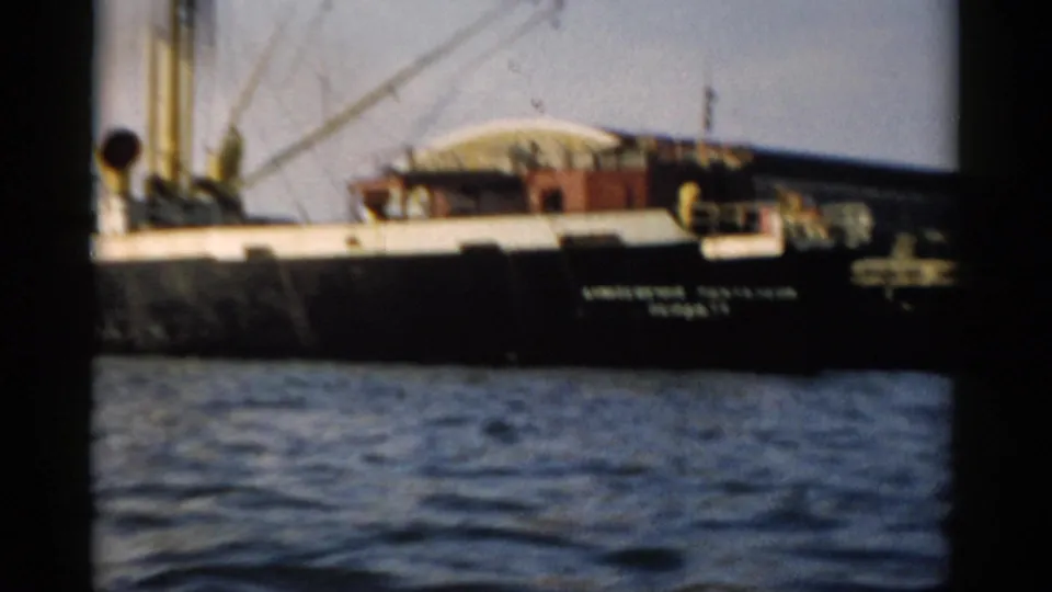 Cargo ship at close range showing what appears to be a Cyrillic hull inscription, red-painted upper deck structures, and derrick crane arms, circa 1957.