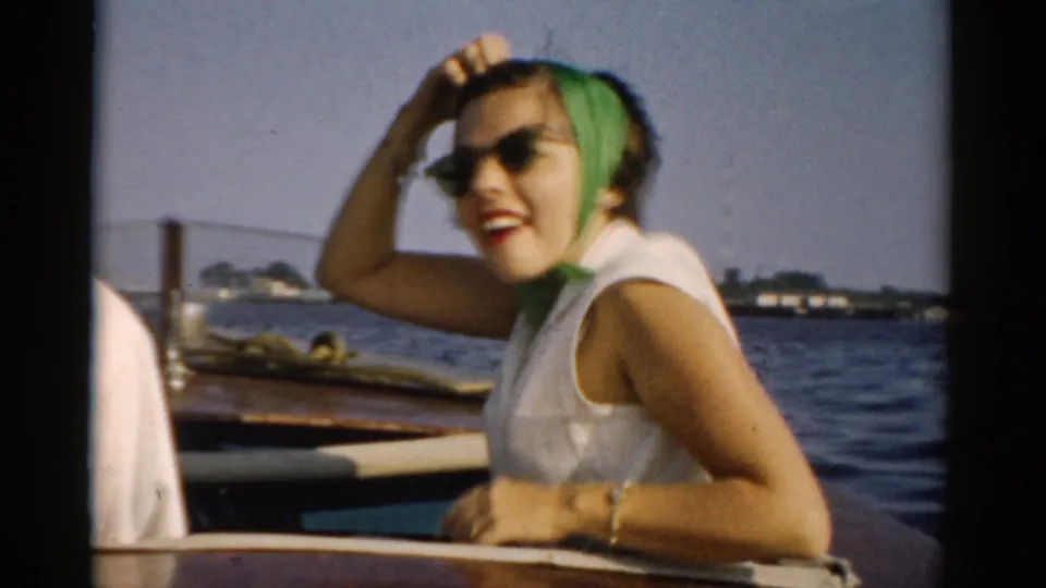 A woman in 1950s leisure attire, white sleeveless top, green headscarf and cat-eye sunglasses, seated on a wooden boat on open water with a distant shoreline behind her, circa 1957.