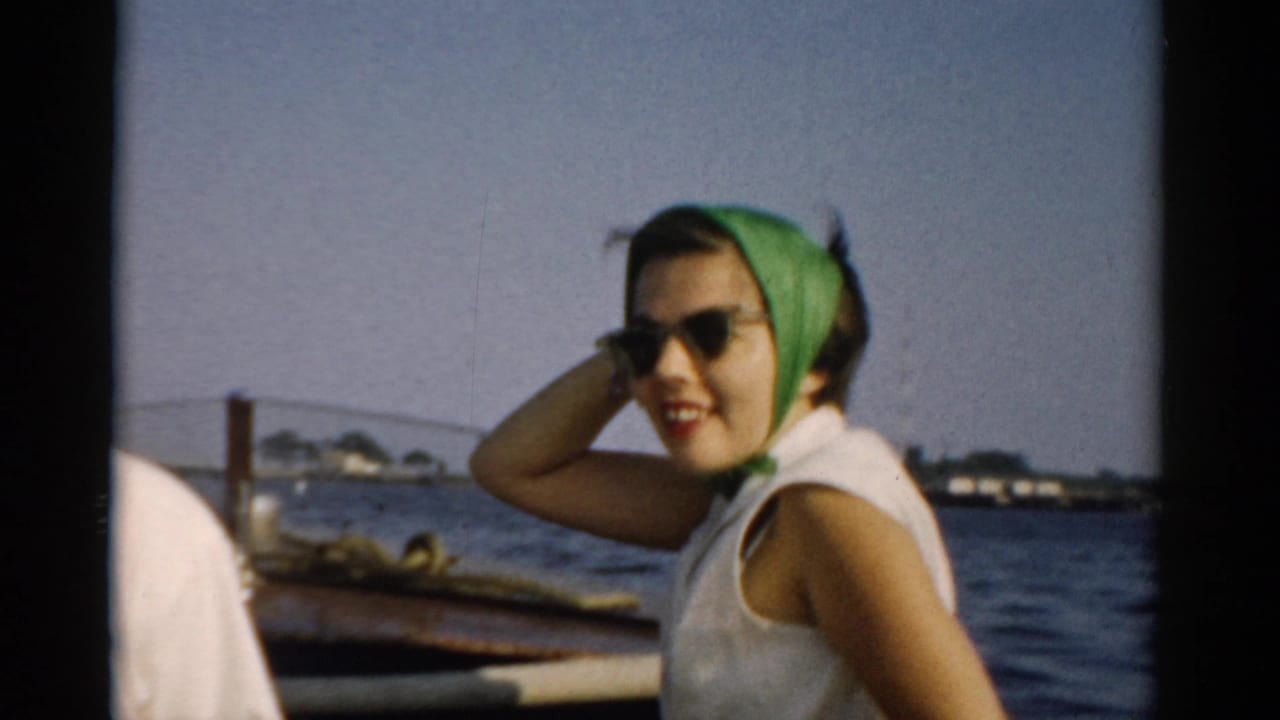 Young woman in green headscarf and cat-eye sunglasses seated in a wooden boat, hand raised near her ear, open water and low shoreline behind.