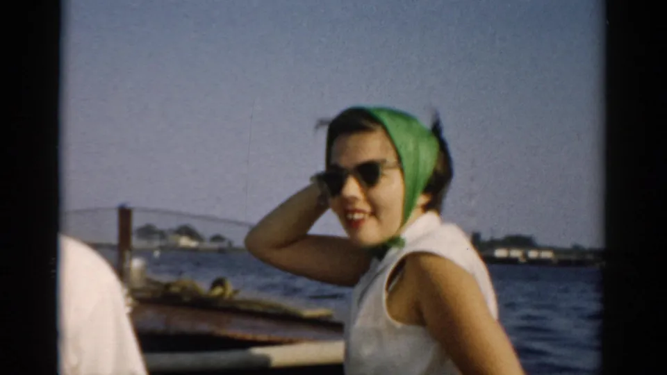 Young woman in late-1950s casual dress (green headscarf, white sleeveless top, cat-eye sunglasses) posed in a wooden boat on a lake, likely Catskills, New York, circa 1957.