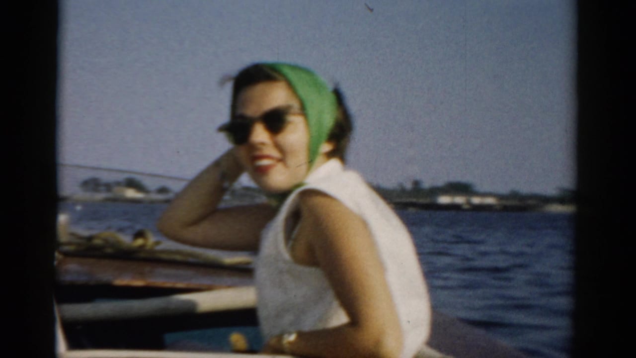 Young woman in green headscarf and cat-eye sunglasses smiling toward camera from a wooden boat; low shoreline with structures visible in the background, circa 1957, Catskills region, New York.