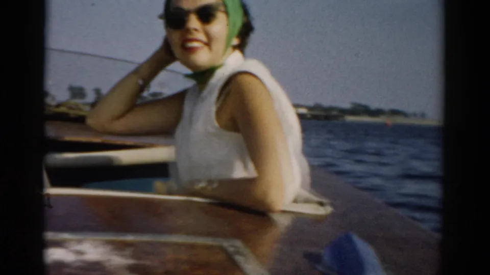 Young woman in green headscarf and cat-eye sunglasses facing camera with a broad smile; wooden boat interior in foreground and open water with distant shoreline behind, circa 1957, Catskills region.