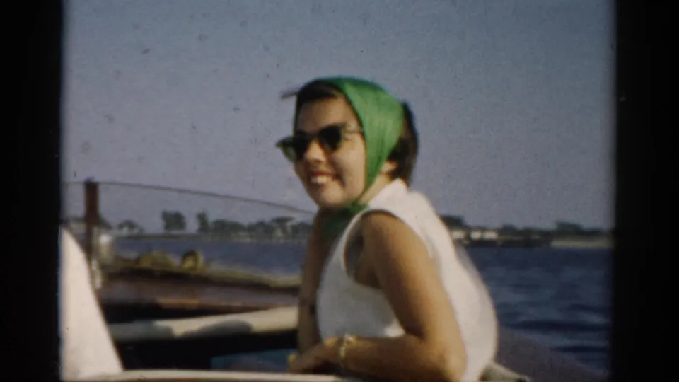 Young woman in green headscarf and cat-eye sunglasses smiling from a wooden boat; a dock piling and low shoreline are visible in the background, circa 1957, Catskills region, New York.