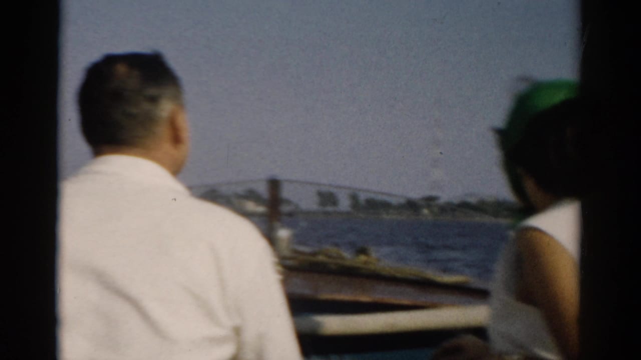 Two passengers in a small motorboat on a harbour, possibly Catskill area, New York, ca. 1957; man seen from behind, woman in green headscarf alongside.
