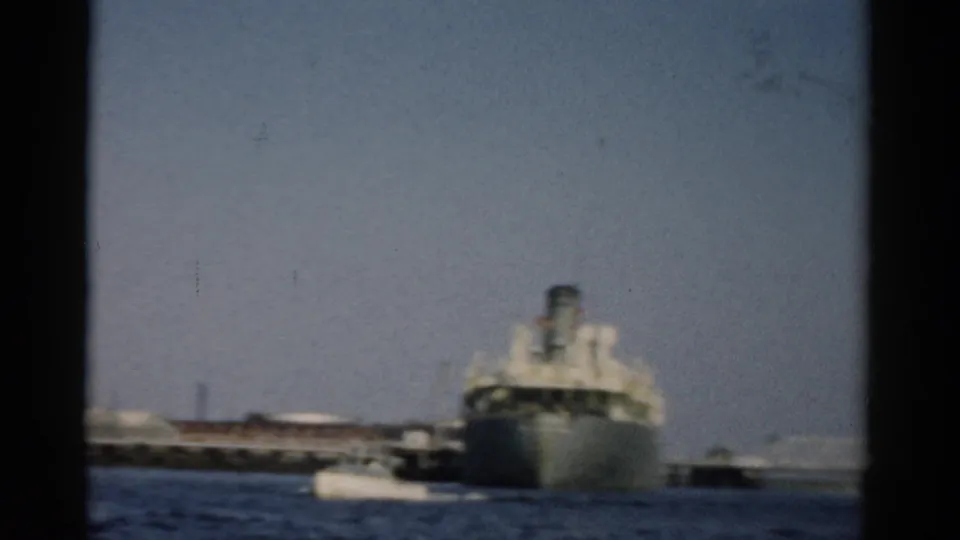 Mid-century ship on open water filmed from a small boat, industrial port structures visible in background, circa 1957 home movie.