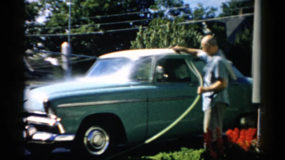 Adult male hosing a two-tone light-blue 1950s American sedan in a residential driveway, with utility wires overhead and trees behind, filmed on 8mm circa 1957 in the Catskill, New York area.