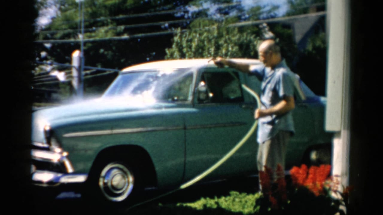 Older man washing a mid-1950s teal two-tone American sedan with a garden hose on a sunny suburban driveway; power lines and trees in background, Catskill, New York, circa 1957.