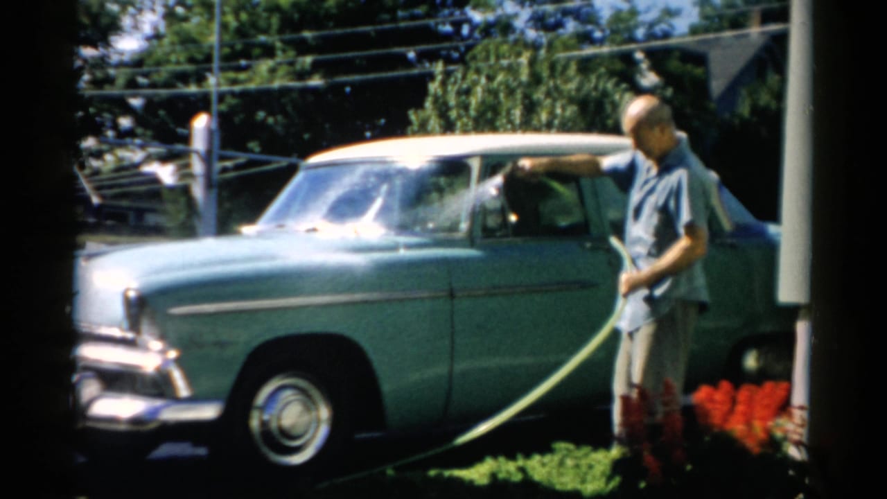 Older man rinsing the windshield of a mid-1950s teal two-tone sedan with a garden hose on a bright sunny driveway; Catskill, New York, circa 1957.
