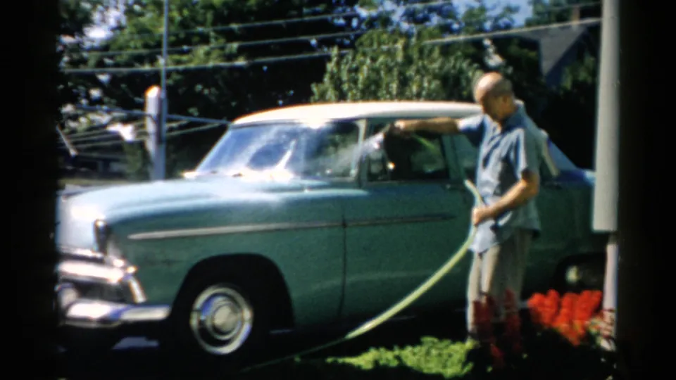 Older man rinsing the windshield of a mid-1950s teal two-tone sedan with a garden hose on a bright sunny driveway; Catskill, New York, circa 1957.