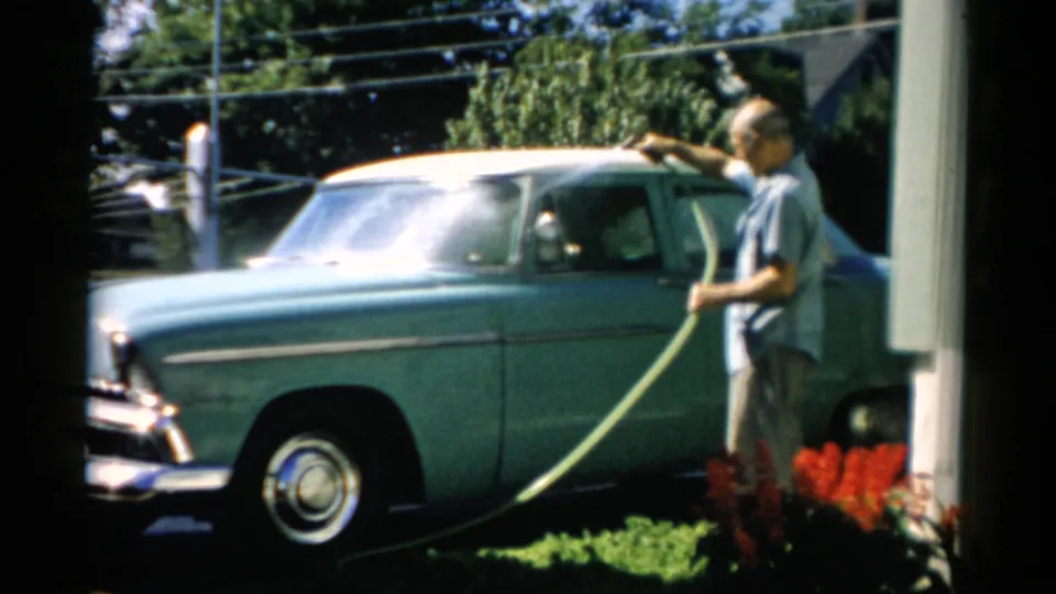 Man pausing mid-wash beside a mid-1950s teal American sedan, chrome grille prominent in foreground; red flowers border the driveway, Catskill, New York, circa 1957.