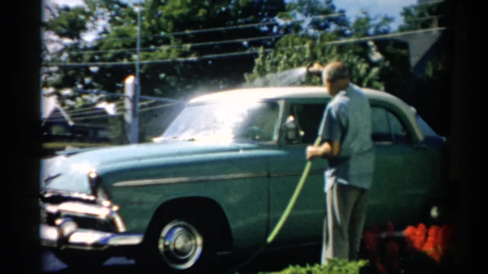 Older man hosing down the front hood and grille area of a mid-1950s teal American sedan on a sunny residential driveway; Catskill, New York, circa 1957.