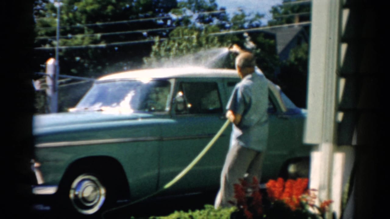 Man in blue shirt hosing a late-1950s American sedan in a home driveway, Catskill NY, 1957; red garden flowers and utility pole visible in background.
