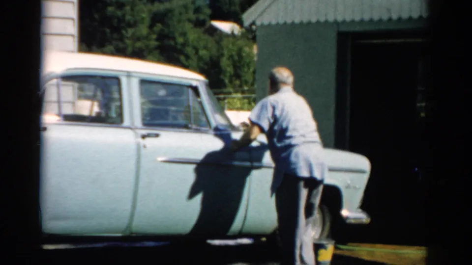Man in blue shirt hand-drying the side of a pale blue late-1950s sedan beside a corrugated metal garage, Catskill NY area, 1957.