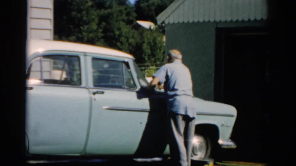 Man in blue shirt attending to the rear of a late-1950s American sedan in a residential driveway beside a corrugated metal garage, Catskill NY, 1957.