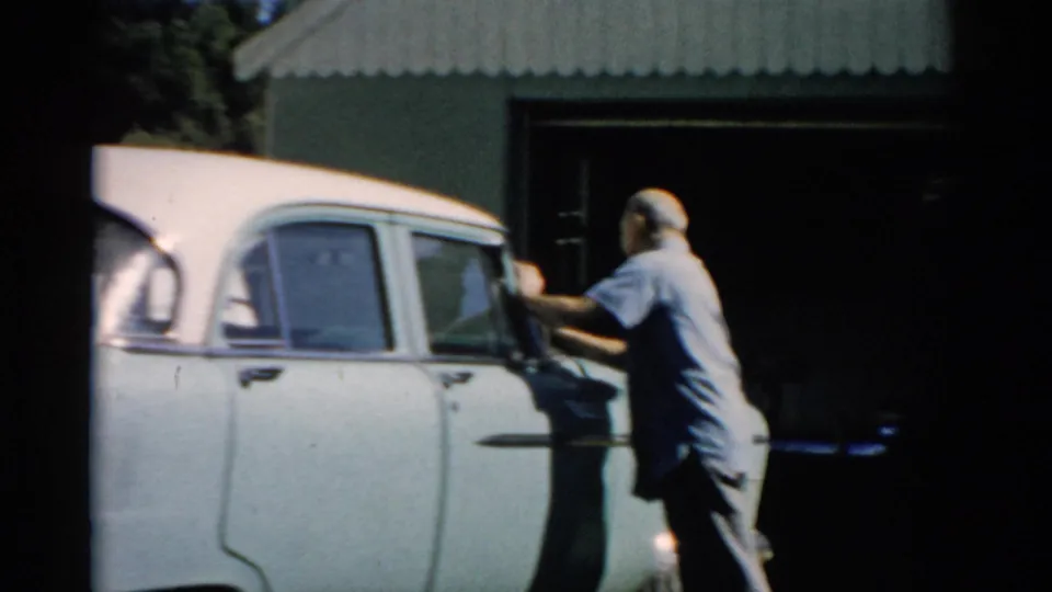 Man in blue shirt wiping the rear door and window of a pale blue late-1950s sedan in a residential driveway, Catskill NY area, 1957.