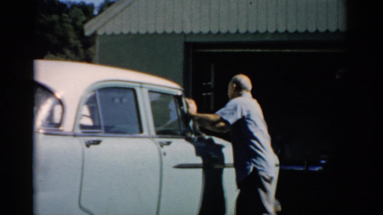 Older man in light blue shirt working on the upper door and window area of a white 1950s sedan in front of a corrugated metal shed.