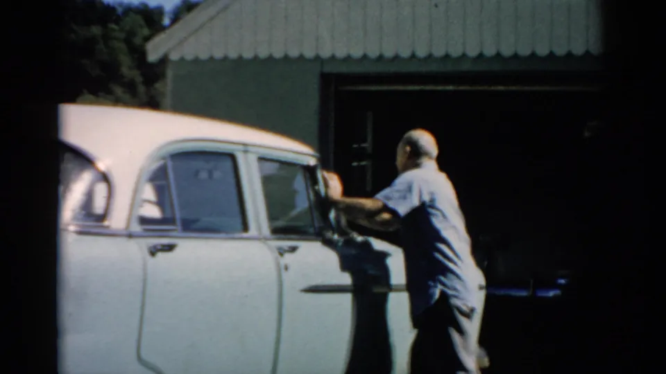 Older man in light blue shirt tending to the upper door or window of a white late-1950s American sedan beside a corrugated metal outbuilding, Catskill, New York, circa 1957.