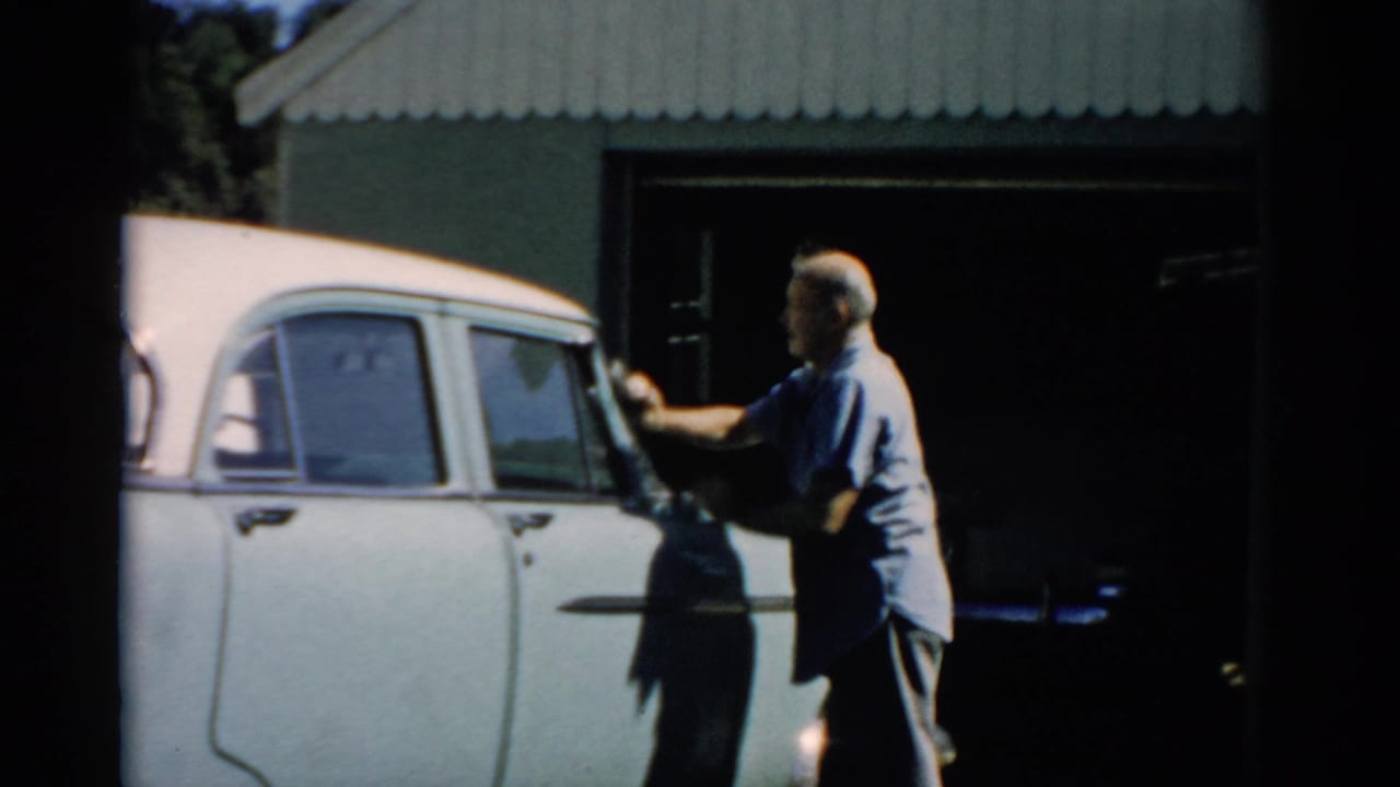 Older man in light blue shirt working along the side of a white late-1950s sedan in profile, corrugated metal shed and open bay behind, Catskill, New York, circa 1957.