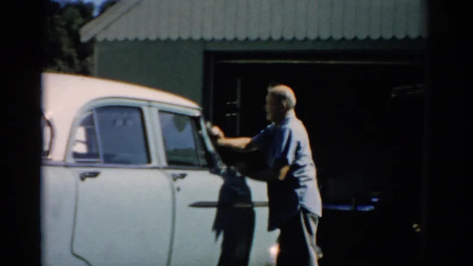 Older man in light blue shirt working along the side of a white late-1950s sedan in profile, corrugated metal shed and open bay behind, Catskill, New York, circa 1957.