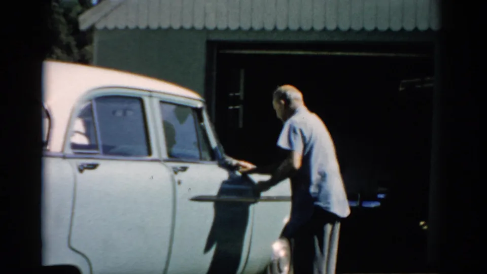 Older man working on the lower body of a white late-1950s sedan with a dark object (possibly a hose or bucket) at his feet, corrugated metal outbuilding behind, Catskill, New York, circa 1957.