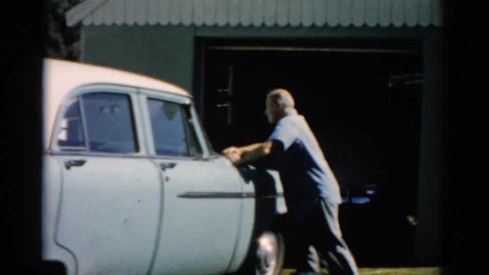 Older man wiping or pressing his hand on the door panel of a white late-1950s sedan in bright summer light, corrugated metal outbuilding behind, Catskill, New York, circa 1957.