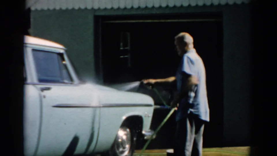 Man hosing down a pale blue mid-1950s American sedan in front of a corrugated-roof garage, Catskill, New York, circa 1957.