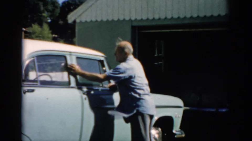 Man cleaning the rear door area of a pale blue mid-1950s sedan, dark garage and corrugated roof behind, circa 1957.