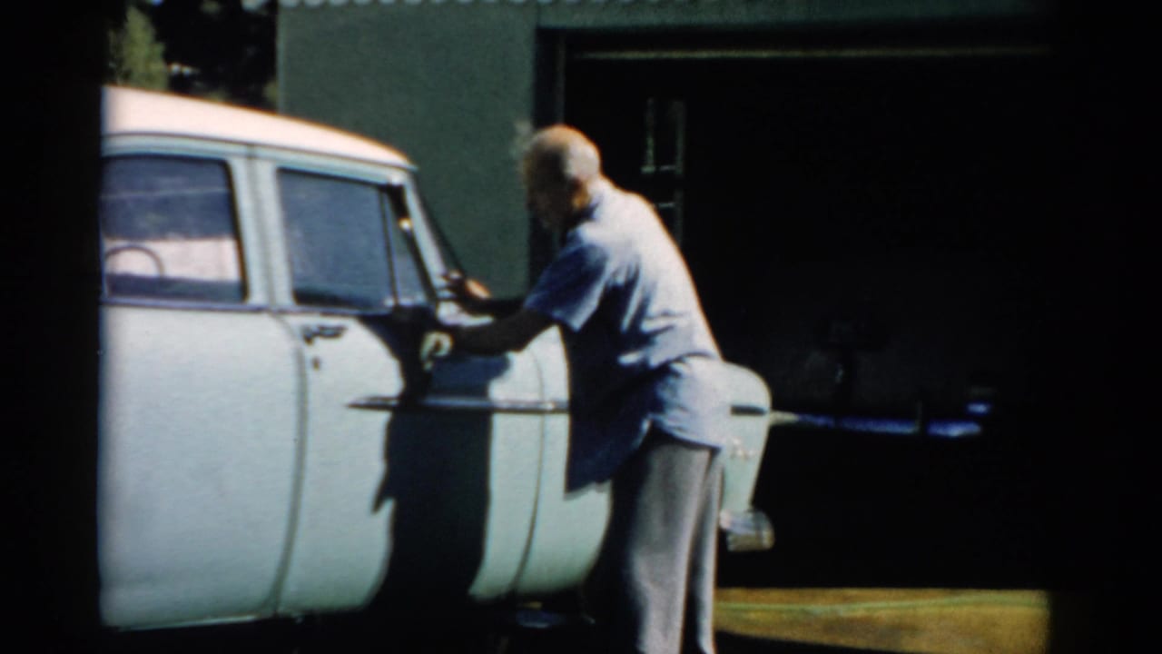 Older bald man in a blue-grey shirt wiping the driver's side of a pale station wagon parked in front of a dark green garage.