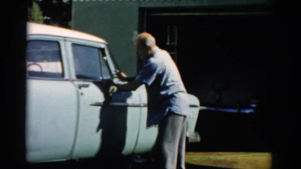 Older bald man hand-wiping the driver's-side panel of a late-1950s pale station wagon, dark green corrugated garage behind, 8mm home-movie footage, Catskill, New York, c. 1957.