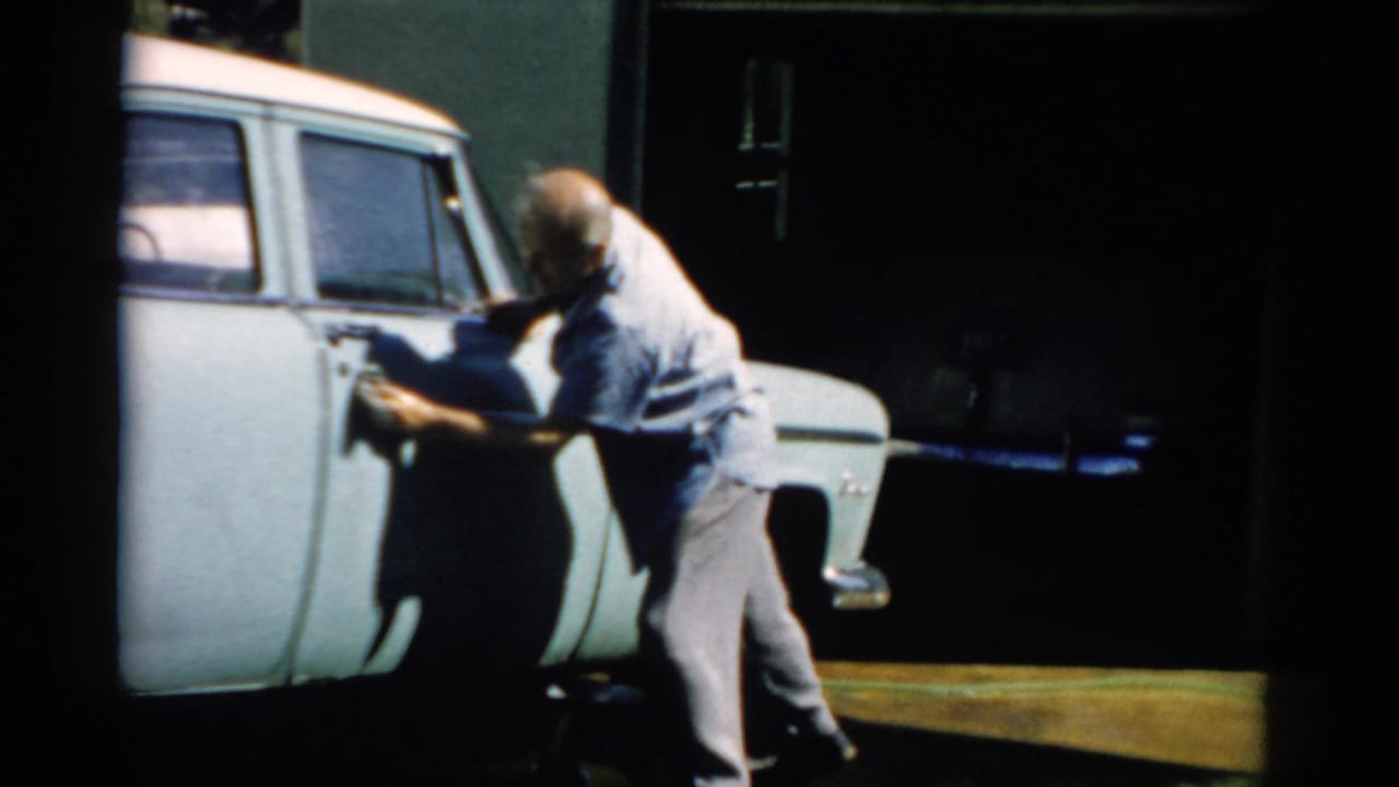 Older bald man leaning toward the front door area of a pale station wagon, large side windows and green building visible behind.