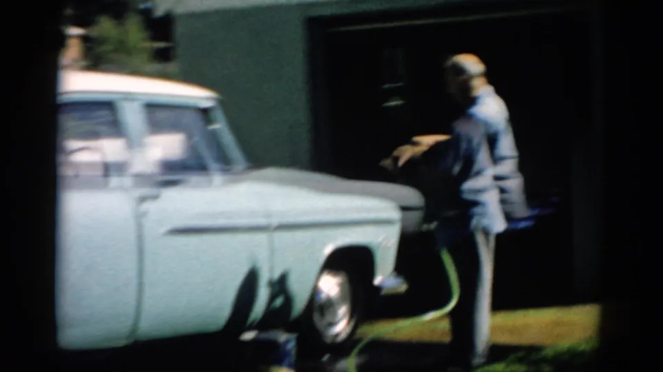 Older bald man moving toward the rear of a late-1950s pale station wagon, green garden hose coiled on driveway, chrome hubcap visible, Catskill, New York, c. 1957.