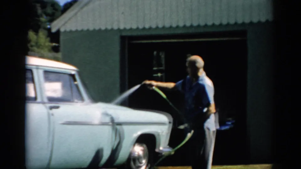 Older bald man hosing down the front end of a late-1950s pale station wagon, green hose clearly visible, corrugated metal garage and summer trees behind, Catskill, New York, c. 1957.
