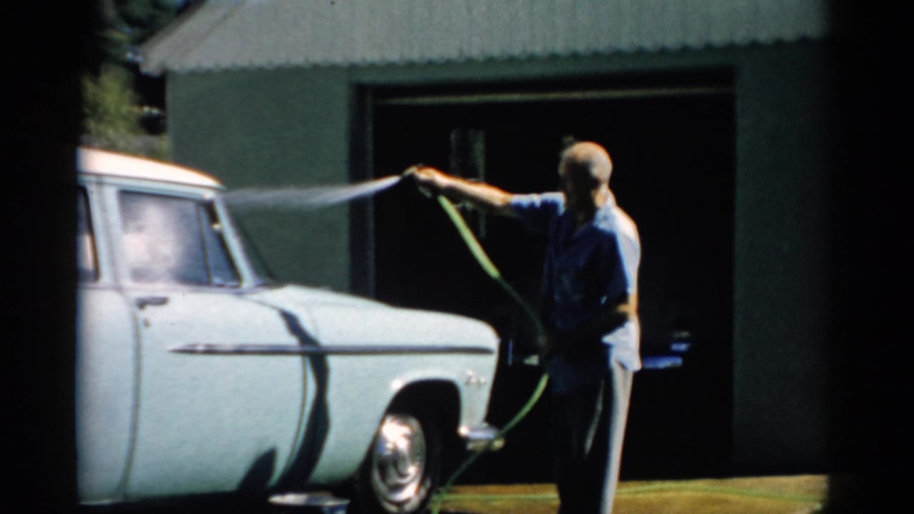 Man hosing down a pale 1950s American sedan in front of a dark green corrugated metal garage.