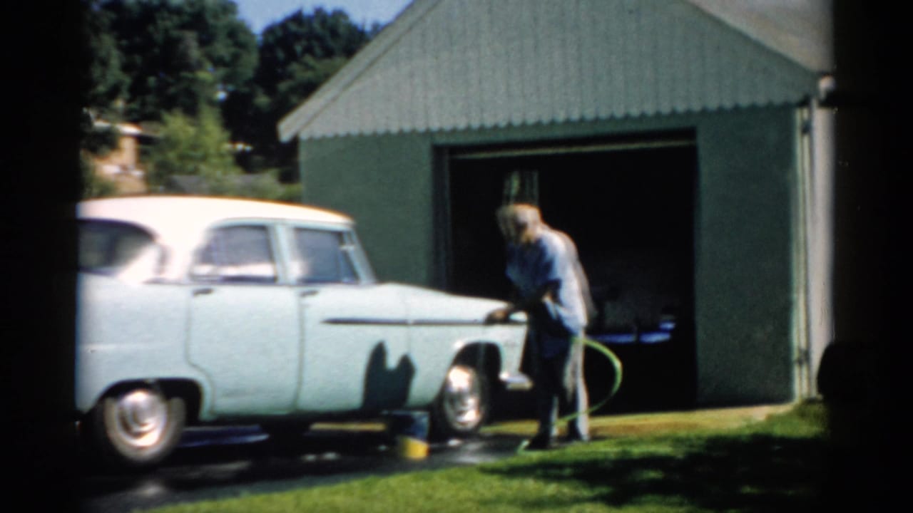 Full view of a pale blue-white 1950s American sedan with chrome hubcaps in a driveway; man crouched at rear with hose, dark green corrugated garage behind, circa 1957 Catskill, New York.