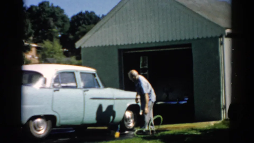 Man crouches to hose the front wheel well of a pale 1950s American sedan outside a corrugated green metal garage, likely Catskill, New York, circa 1957.