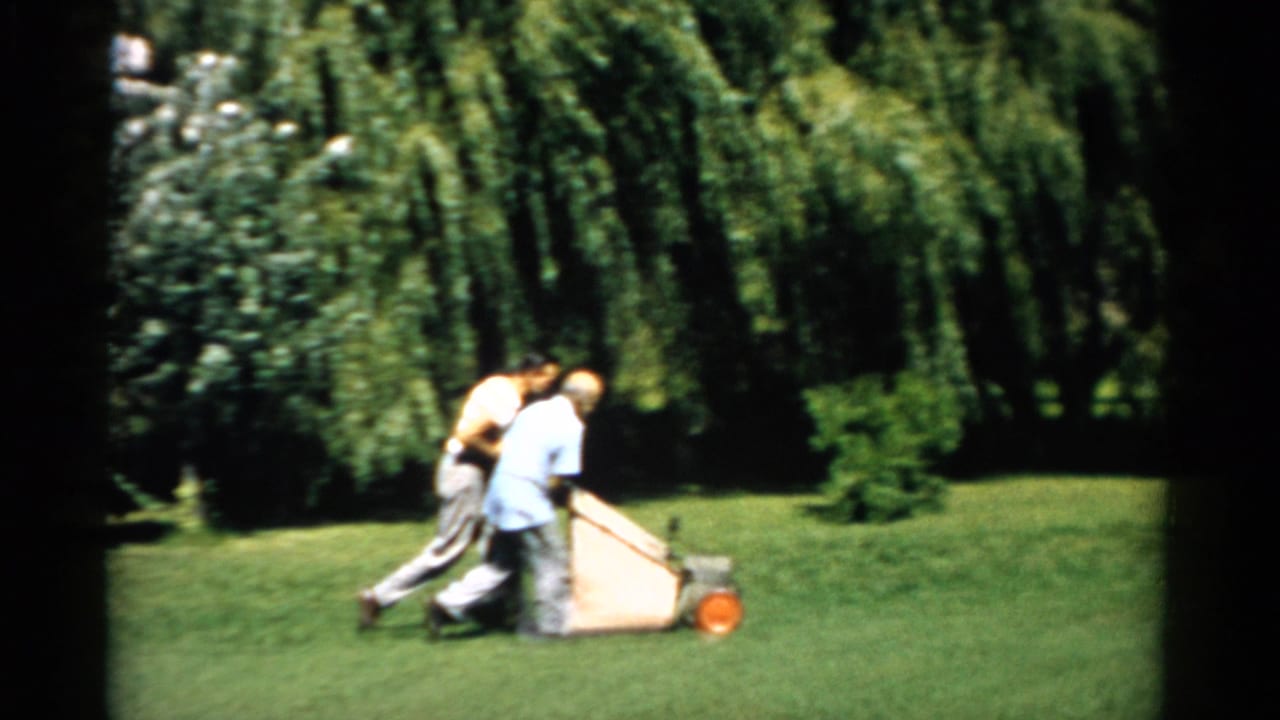Two men pushing a manual push-reel mower with grass catcher on a well-kept lawn in Catskill, New York, circa 1957; weeping willow trees visible behind.